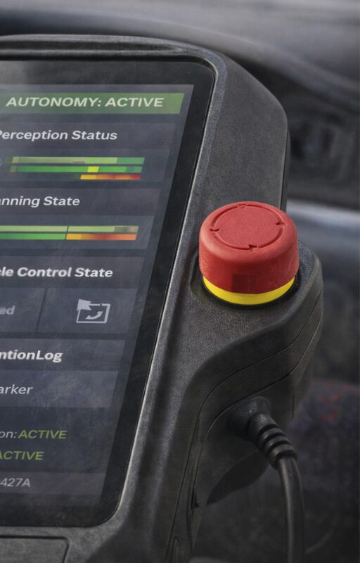 Close-up of a teach pendant showing a touchscreen interface with autonomy status indicators and a red emergency stop pushbutton mounted on the side of the housing.